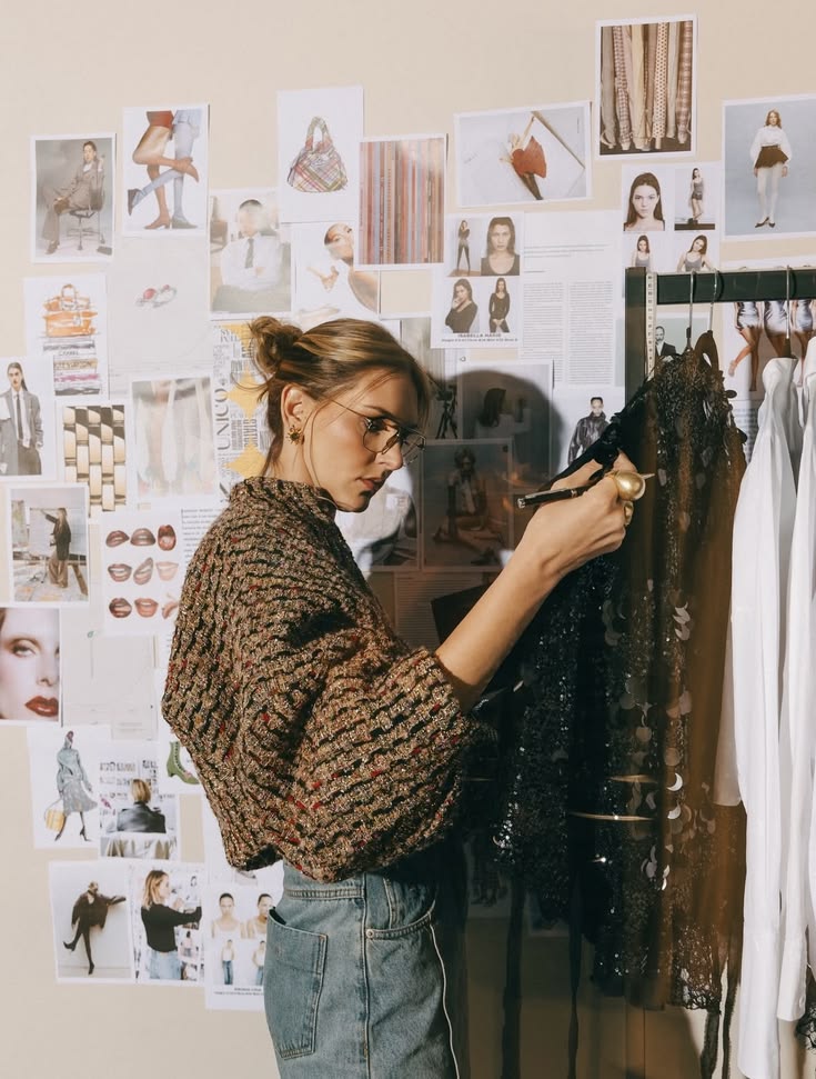 A stylist in glasses pins a sequinned piece on a clothes rail in front of a wall of fashion tear sheets